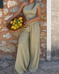 A woman wearing a sage green one-shoulder jumpsuit stands against a rustic stone wall, holding a wooden bowl filled with lemons.