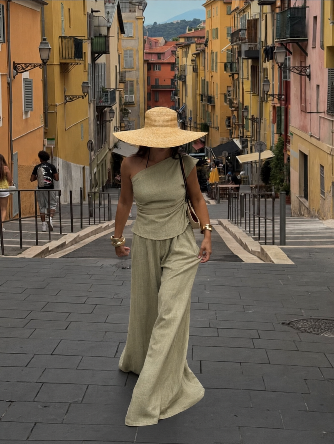 A woman wearing a wide-brimmed straw hat and a matching beige one-shoulder top and wide-leg trousers walks down a cobblestone street lined with colorful yellow and orange buildings.
