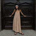 A woman wearing a beige maxi dress and sunglasses stands in front of a large dark wooden door, posing with her hands on the doorframe.
