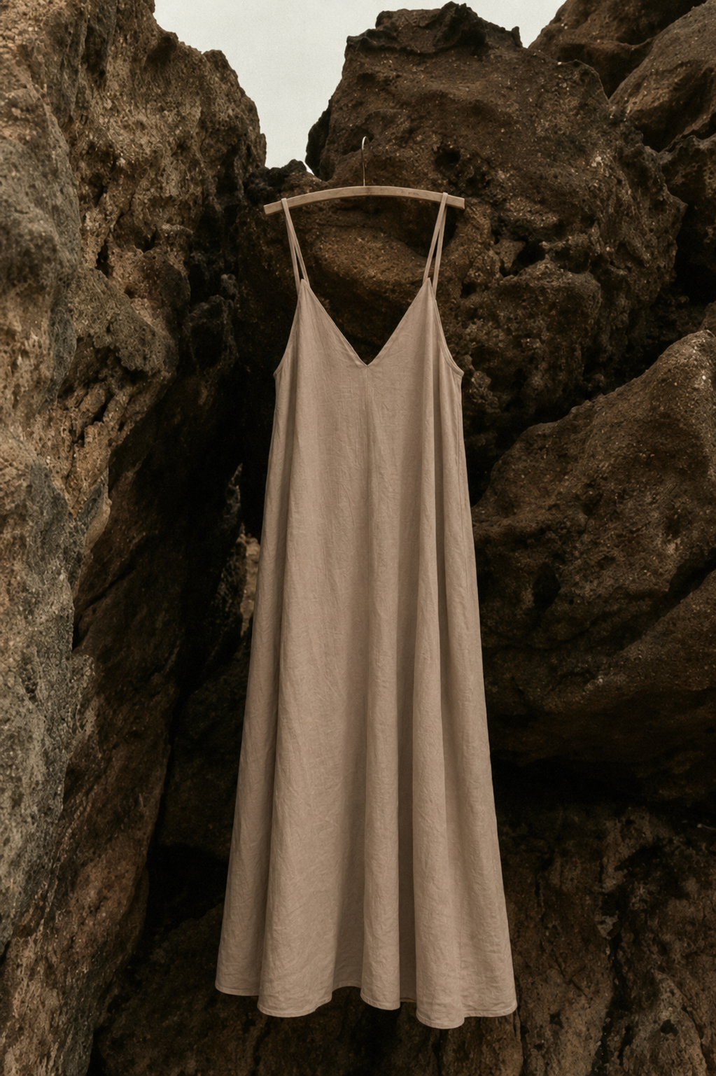A sleeveless beige slip dress with thin straps hangs on a wooden hanger against a backdrop of dark, textured rocks.