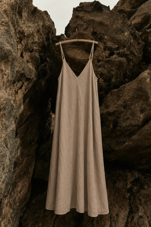 A sleeveless beige slip dress with thin straps hangs on a wooden hanger against a backdrop of dark, textured rocks.