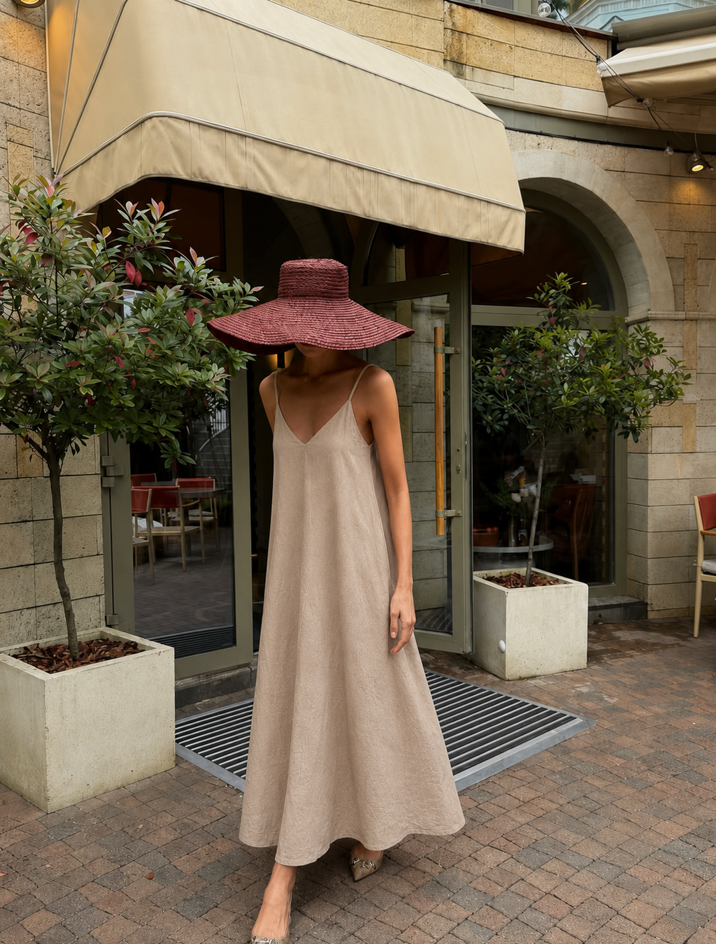 A woman wearing a beige spaghetti-strap maxi dress and a wide-brimmed burgundy hat stands on a brick patio in front of a cafe entrance with stone walls and potted plants.