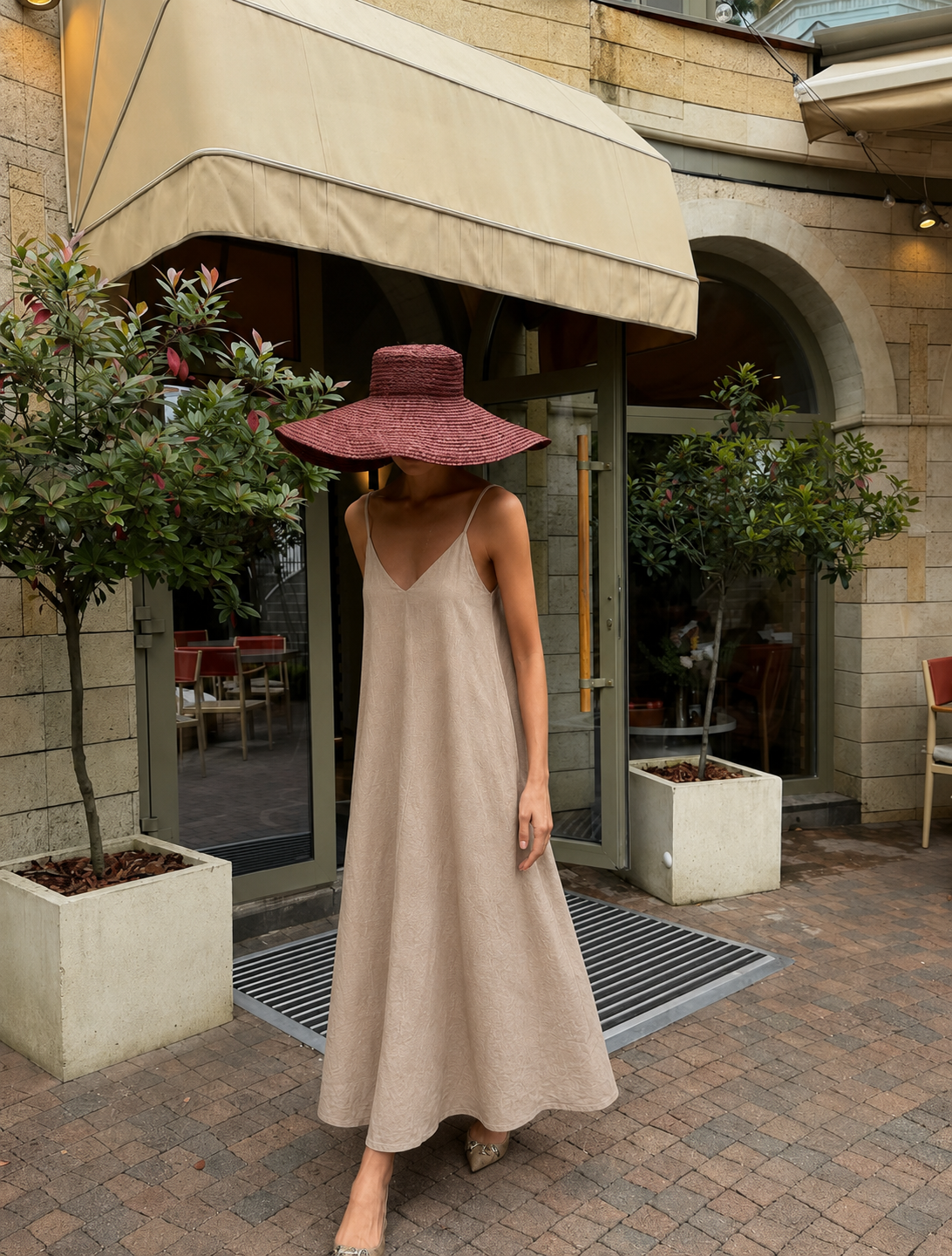 A woman wearing a beige spaghetti-strap maxi dress and a wide-brimmed burgundy hat stands on a brick patio in front of a cafe entrance with stone walls and potted plants.
