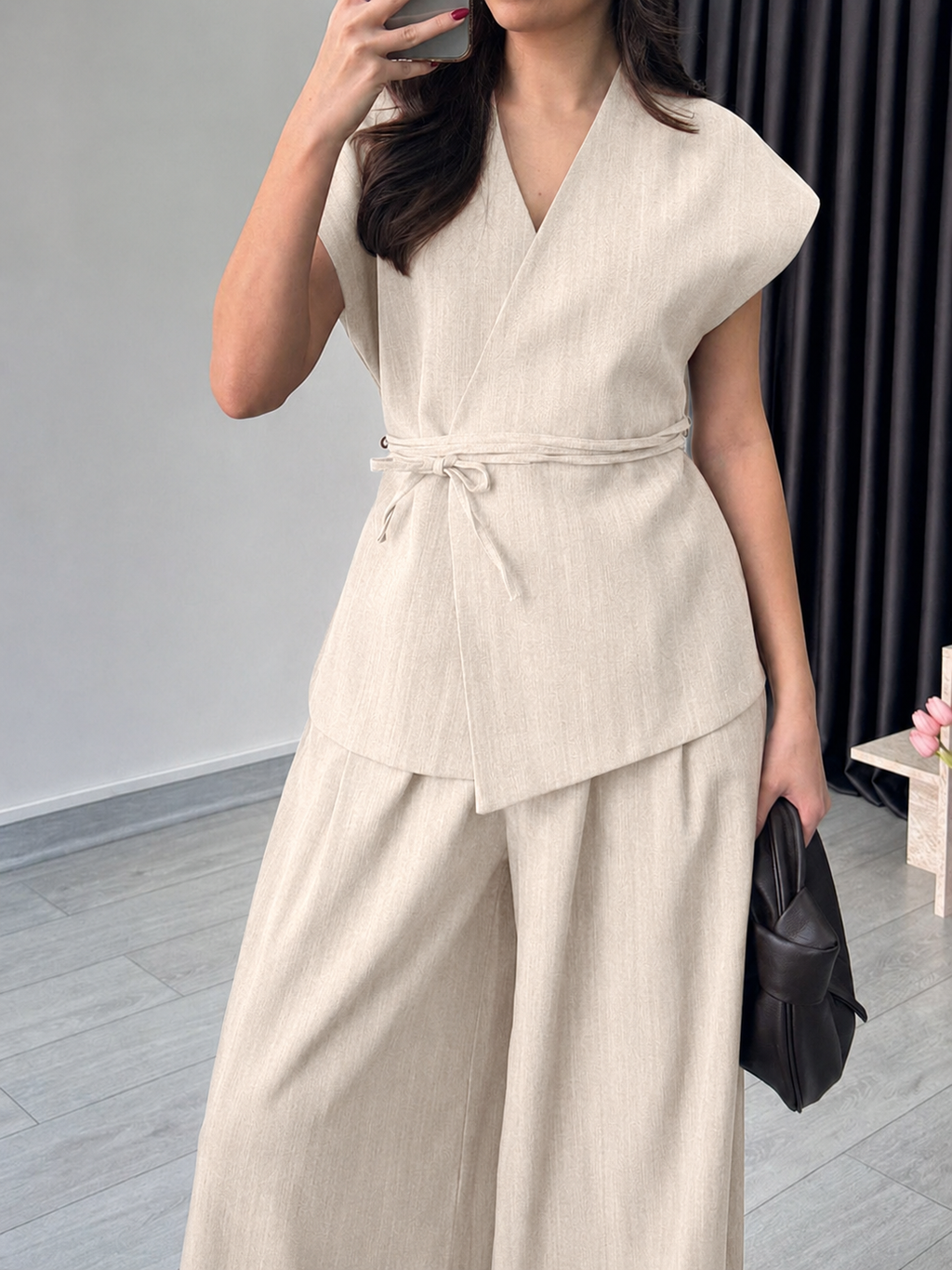 A woman wearing a light beige linen two-piece set consisting of a sleeveless wrap vest with a tie waist and matching wide-leg pants, holding a black handbag.