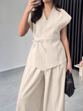 A woman wearing a light beige linen two-piece set consisting of a sleeveless wrap vest with a tie waist and matching wide-leg pants, holding a black handbag.